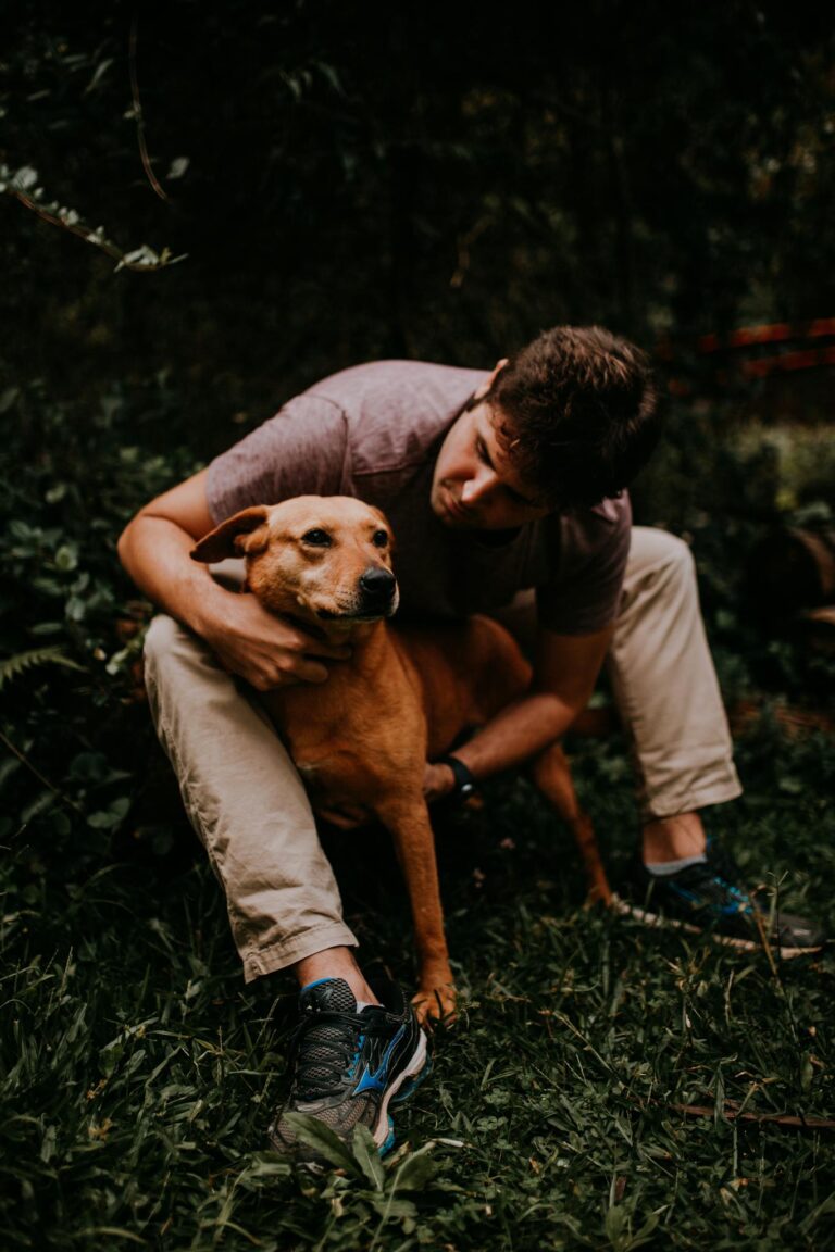 A man lovingly embraces a dog while sitting outdoors in a lush, forested area.