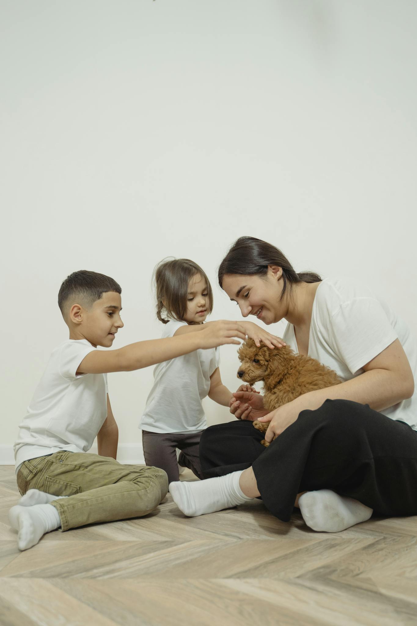 Mother and two children enjoying time with their puppy indoors, sitting on the wooden floor.