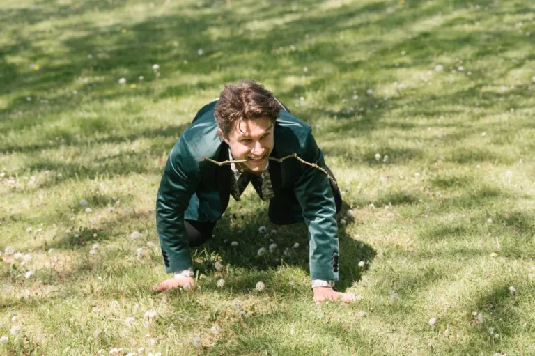 Ein Mann in einer grünen Samtjacke kniet auf allen Vieren im Gras und hält einen kleinen Zweig im Mund, ähnlich wie ein Hund.